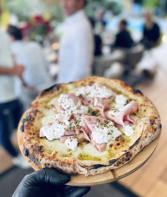 Eine Pizza mit Mortadella, Stracciatella und Pistazien wird auf dem Holzbrett an den Tisch getragen